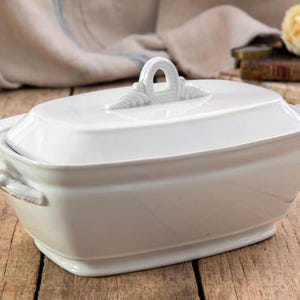 May include: A rectangular white ceramic tureen with a matching lid. The lid features a central arched handle with decorative leaf-like detailing at the base. The tureen has two small, curved handles on the sides and a smooth, glossy finish.