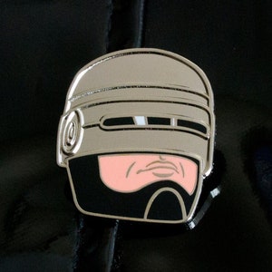 以下が含まれることがあります： 映画「ロボコップ」のキャラクター、ロボコップのヘルメットをあしらったシルバーカラーのエナメルピン。黒、ピンク、シルバーのアクセントが施された詳細なデザインで、SF映画のキャラクターの象徴的な外観を捉えています。