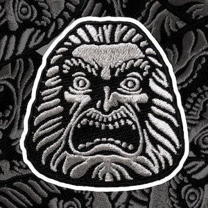 Könnte beinhalten: Ein gestickter Patch in Schwarzweiß, der eine Kreatur mit einem großen Maul und scharfen Zähnen zeigt. Die Augen der Kreatur sind groß und starren. Der Patch ist von einem weißen Rand umgeben.