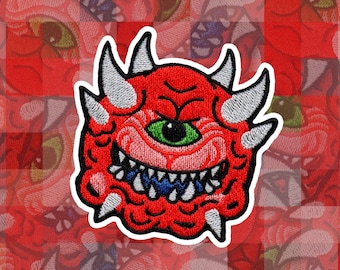 Könnte beinhalten: Roter und weißer gestickter Aufnäher eines einäugigen Monsters mit scharfen Zähnen und Hörnern. Das Monster hat ein grünes Auge und einen blauen Mund.