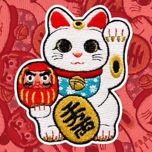 May include: White and red embroidered lucky cat with a raised paw, holding a red and gold daruma doll. The cat is wearing a blue and white collar with a bell and has a gold coin on its chest with the word "MANEKI" written on it.