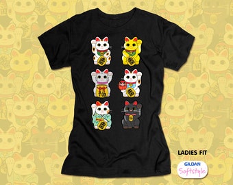 May include: Black t-shirt featuring a design of eight different Maneki-neko cats, each with unique colors and accessories. The text "LADIES FIT" and "GILDAN Softstyle" are printed on the lower right.