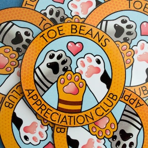 May include: Four colourful round stickers with orange borders and a white background. Each sticker features a cartoon cat paw print design with the text "Toe Beans Appreciation Club" and "Toe Beans" in a yellow and orange font.