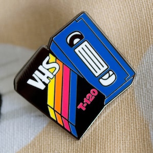 May include: A black and blue VHS tape pin with rainbow stripes and the text "VHS" and "T-120".