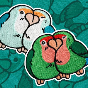 May include: Two pairs of embroidered lovebirds, one pair is blue and white, the other is green and pink. The birds are facing each other and their beaks are touching.