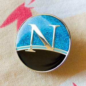May include: A round, blue and black pin with a silver border. The pin features a large, silver letter "N" on a blue background with glitter. The bottom half of the pin is black.
