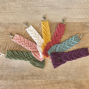 May include: Seven colorful macrame keychains with fringe tassels. The keychains are arranged in a circle on a wooden surface.