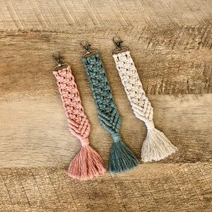 May include: Three macrame keychains with tassels. The keychains are in pink, green, and white.