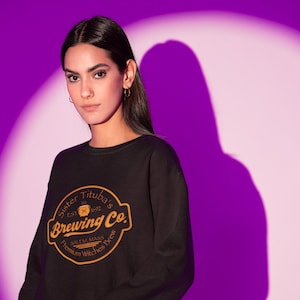 May include: Black sweatshirt with a circular logo that reads "Sister Tituba's Brewing Co." in gold lettering. The logo includes the text "Salem, Mass" and "Premium Witches Brew." The sweatshirt is a crew neck style.