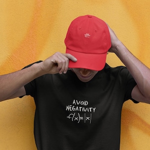 Puede incluir: Una persona con una camiseta negra con el texto "AVOID NEGATIVITY f(x)=|x|" y una gorra de béisbol roja.