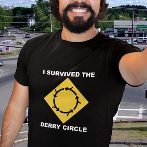 May include: Black t-shirt with the text "I Survived the Derry Circle" and a yellow and black graphic. The graphic is a yellow diamond with a circular arrow design. The person in the image is wearing a blue baseball cap.