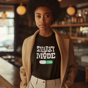 May include: A woman wearing a brown jacket over a black t-shirt with the text "SNARKY MODE ON" in white. The t-shirt has a white toggle switch with a green "ON" indicator.