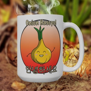 Könnte beinhalten: Eine weiße Keramiktasse mit einer Cartoon-Illustration einer Zwiebel und dem Text "Onion Haseyo!" in Englisch und Koreanisch. Die Zwiebel lächelt und hat Arme und Beine.