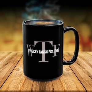 Op de afbeelding: Zwarte koffiemok met witte tekst "Whiskey Tango Foxtrot" op een houten oppervlak.