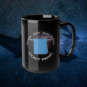 Könnte beinhalten: Schwarze Keramik-Kaffeetasse mit einem weißen Kreis-Motiv, das ein blaues Handtuch zeigt, das an einem Handtuchhalter hängt, einen Galaxie-Hintergrund und den Text "Towel Day May 25th Don't Panic".