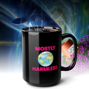 Könnte beinhalten: Eine schwarze Keramik-Kaffeetasse mit den rosa Worten "MOSTLY HARMLESS" und einem Bild der Erde. Die Tasse ist mit einer dunklen Flüssigkeit, möglicherweise Kaffee, gefüllt, und Dampf steigt auf. Ein kleiner Blumentopf ist im Hintergrund zu sehen.