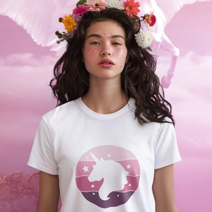 Op de afbeelding: Een wit T-shirt met een roze en paarse ombre cirkelvormig ontwerp met een wit silhouet van een eenhoorn. Het shirt wordt gedragen door een persoon met lang bruin haar en een bloemenkrans.