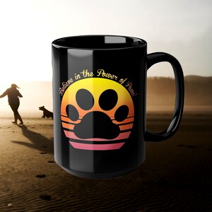 May include: Black ceramic mug featuring a sunset-coloured paw print design and the text "Believe in the Power of Paws!"