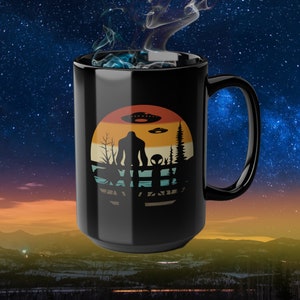 May include: Black ceramic coffee mug with a retro-style design featuring a silhouette of a Bigfoot-like creature, a small alien, and flying saucers against a sunset background.