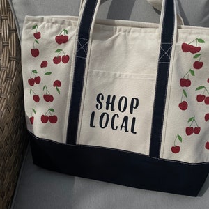 Może przedstawiać: Biała torba typu tote z pł&oacute;tna z czarnymi paskami i czarnym dnem. Torba ma kieszeń z przodu z nadrukiem "SHOP LOCAL" w kolorze zielonym. Torba jest ozdobiona czerwonymi wiśniami na białym tle.