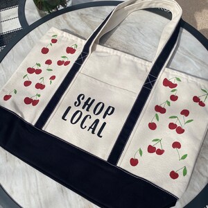 Może przedstawiać: Biała torba typu tote z pł&oacute;tna z czarnymi paskami i czarnym dnem. Torba ma nadruk czerwonych wiśni i napis "SHOP LOCAL" w kolorze czarnym.
