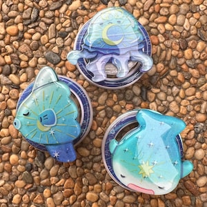 May include: Three colorful phone grips with celestial designs. The grips feature a blue jellyfish, a blue and yellow sun, and a blue whale, all with a glossy finish. Each grip is mounted on a circular base with a blue and white pattern.