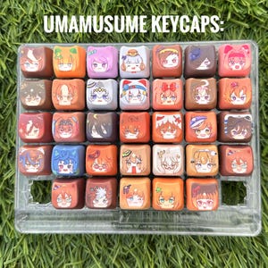 Könnte beinhalten: Eine Sammlung von vierunddreißig quadratischen Tastenkappen in einer durchsichtigen Kunststoffschale, jede mit einem einzigartigen, farbenfrohen Anime-Gesicht. Der Text UMAMUSUME KEYCAPS ist oben über dem Raster der Tastenkappen aufgedruckt.