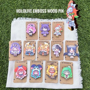 Op de afbeelding: Een collectie van twaalf Hololive houten pins met diverse anime-stijl personages in kleurrijke outfits. Elke pin is bevestigd op een rechthoekige bruine kaart met het label 3D WOODPIN. De pins tonen uiteenlopende haarkleuren en kostuums.