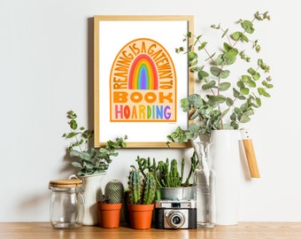 Könnte beinhalten: Ein gerahmter Druck mit einem Regenbogenbogen und dem Text "Reading is a gateway to book hoarding". Der Druck ist auf weißem Hintergrund und von Pflanzen und einer weißen Kanne umgeben.