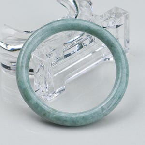 Könnte beinhalten: Ein runder, hellgrüner Jade-Armreif. Der Armreif ist ein massives, glattes Band mit leicht durchscheinendem Aussehen. Der Jade hat subtile Farbvariationen mit Andeutungen von dunklerem Grün. Der Armreif wird auf einem klaren, geometrischen Ständer präsentiert.