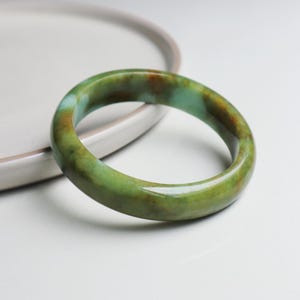 May include: A jade bangle bracelet in shades of green, brown, and turquoise. The polished surface of the bracelet reflects light, showing the natural colour variations. The bracelet is circular and designed to be worn on the wrist.