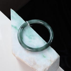 May include: A smooth, circular jade bangle bracelet featuring a translucent, deep forest green colour with subtle darker green inclusions and a polished, reflective surface. A small white string tag is attached to the top of the bangle.