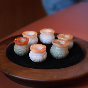 May include: Six small, decorative jade pots with a flower-like rim. The pots are white, orange, and green, with carved swirl designs on the base. They are arranged on a black surface within a wooden tray.
