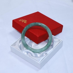 Puede incluir: Un brazalete de jade verde descansa en una caja blanca forrada de satén, con una caja roja detrás. El brazalete es circular y liso. La caja roja es rectangular con un diseño floral dorado en la parte superior. El brazalete mide unos 7 cm de diámetro.