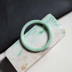 Pode incluir: Uma pulseira circular lisa, feita de jade verde pálido com subtis inclusões manchadas em branco e verde escuro. A pulseira apresenta uma superfície polida e arredondada com uma espessura consistente ao longo de toda a sua circunferência.