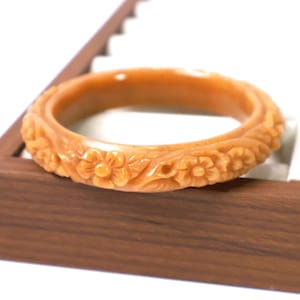 May include: A circular bangle bracelet in a warm, light orange hue, featuring a continuous carved relief pattern of small five-petaled flowers and delicate leaves encircling the entire outer surface of the smooth, polished band.