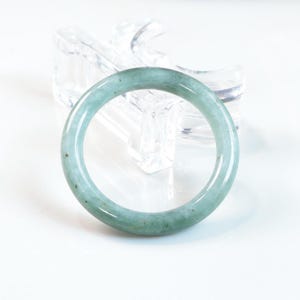 May include: A smooth, circular bangle bracelet crafted from pale green jade stone. The bangle features a polished, glossy finish with subtle natural variations in colour, including faint darker green flecks and translucent areas throughout the stone.
