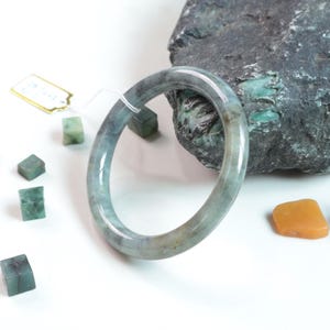 Pode incluir: Uma pulseira de jade lisa e circular com uma paleta de cores verde e cinza translúcida e manchada. A pulseira tem um acabamento polido e brilhante e é acompanhada por uma pequena etiqueta branca e várias peças pequenas de pedra verde quadradas.