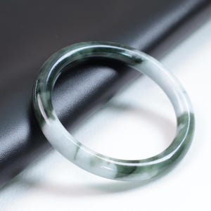 May include: A smooth, polished bangle bracelet crafted from stone, featuring a rounded profile. The surface displays a marbled pattern with translucent white sections transitioning into deep, dark forest green swirls throughout the circular band.
