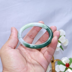 May include: A jade bangle bracelet, displaying a colour gradient from white to deep green. The polished surface reflects light, accentuating the natural stone variations. This elegant piece is a classic jewellery item.