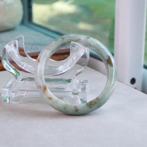 May include: A smooth, circular bangle bracelet crafted from pale green stone with natural, mottled brown inclusions throughout its surface. The bangle is displayed upright, resting within a clear, curved acrylic stand designed for support.