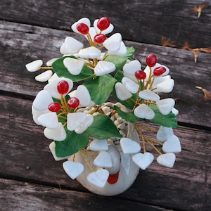 Puede incluir: Arreglo floral decorativo en una maceta color crema. El arreglo presenta piedras blancas en forma de corazón, bayas rojas y hojas verdes. Los tallos son dorados y la maceta tiene un diseño de cara sonriente.