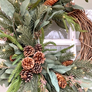 Puede incluir: Una corona artesanal con una base de vid natural, adornada con ramas de hoja perenne artificiales, hojas de eucalipto y varias pi&ntilde;as marrones. La corona tiene una est&eacute;tica r&uacute;stica y natural, perfecta para la decoraci&oacute;n del hogar de temporada.