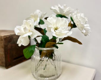 May include: A clear glass vase with vertical ridges holds a bouquet of white artificial flowers. The flowers have large, layered petals and are surrounded by green leaves and brown accents. The vase sits on a white surface.