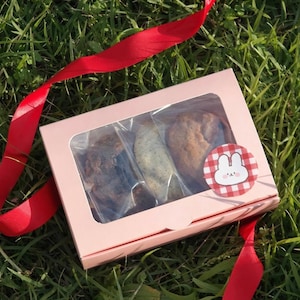 May include: A pink box with a clear window showing three different types of cookies. The box has a white bunny rabbit sticker with red polka dots.