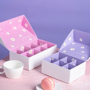 May include: Two white gift boxes with a divided interior for holding small items. The boxes have a white base and a lid with a floral pattern. One box has a pink lid with white flowers and the text "Lucky Day Lucky Life". The other box has a purple lid with white flowers and the text "Lucky Day Lucky Life".