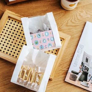 May include: Two white paper bags with clear plastic windows. One bag contains a pink box with a white and blue pattern. The other bag contains a stack of biscuits.