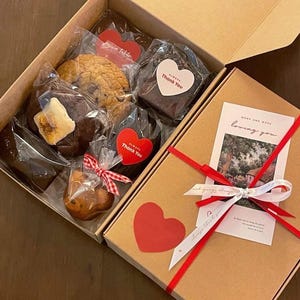 Peut inclure: Une boîte cadeau marron avec un ruban rouge et un ruban blanc. La boîte contient une variété de produits de boulangerie, dont des brownies, des biscuits et un petit gâteau. La boîte contient également une carte avec le texte "More and more loving you".