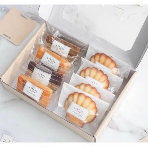 Puede incluir: Una caja blanca llena de pasteles empaquetados individualmente. Los pasteles incluyen madeleines y pasteles rectangulares. El empaque es de pl&aacute;stico transparente con una etiqueta blanca que dice "Bakery Life".