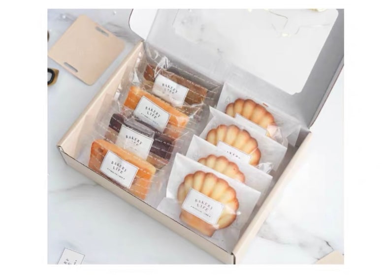 Puede incluir: Una caja blanca llena de pasteles empaquetados individualmente. Los pasteles incluyen madeleines y pasteles rectangulares. El empaque es de pl&aacute;stico transparente con una etiqueta blanca que dice "Bakery Life".